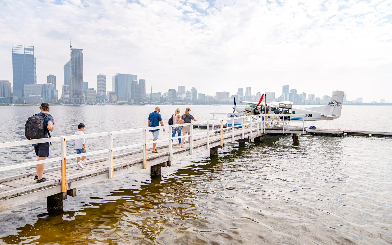 Passengers walking on a jetty towards a seaplane with Perth skyline in the background.
