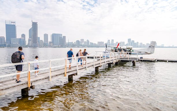 Passengers walking on a jetty towards a seaplane with Perth skyline in the background.