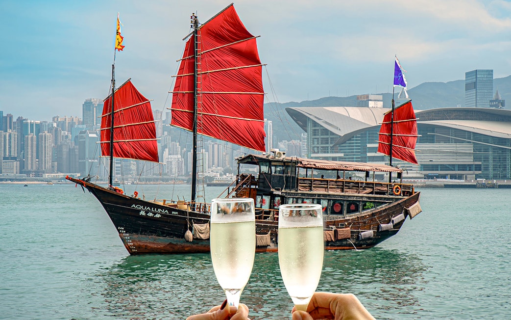 AquaLuna boat with red sails in Victoria Harbour, Hong Kong, with two champagne glasses in foreground.
