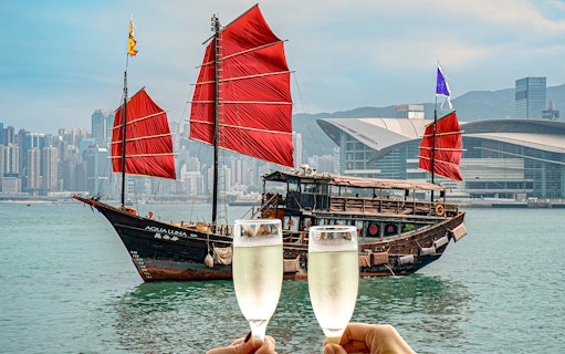 AquaLuna boat with red sails in Victoria Harbour, Hong Kong, with two champagne glasses in foreground.