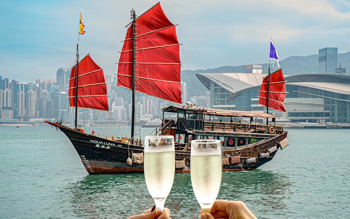 AquaLuna boat with red sails in Victoria Harbour, Hong Kong, with two champagne glasses in foreground.
