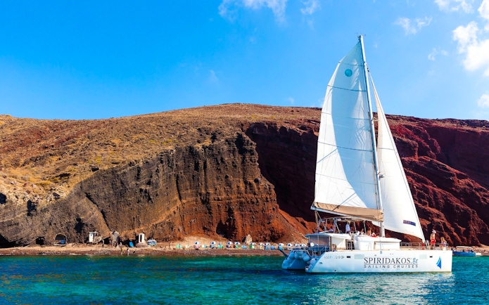Catamaran sailing near the red cliffs of Santorini, Greece.