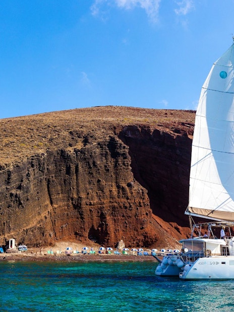 Catamaran sailing near the red cliffs of Santorini, Greece.