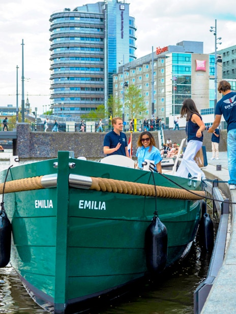 Amsterdam canal cruise boarding with passengers near modern buildings.