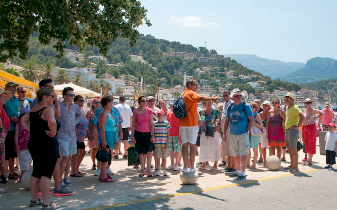 Tour group in Mallorca listening to a guide near the harbor, with mountains in the background.