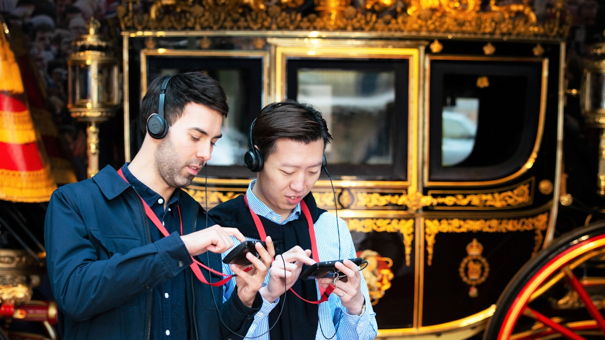 Two men using audio guides in front of a royal carriage at Buckingham Palace.