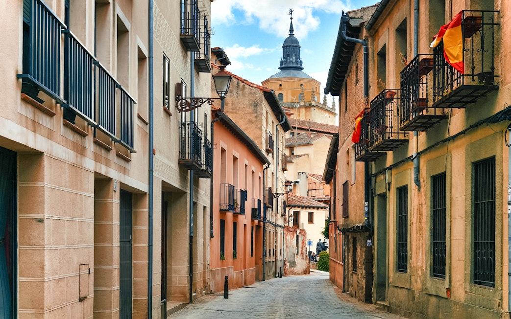 Medieval street in Segovia with historic buildings and a distant church tower.