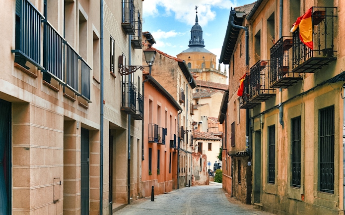 Medieval street in Segovia with historic buildings and a distant church tower.