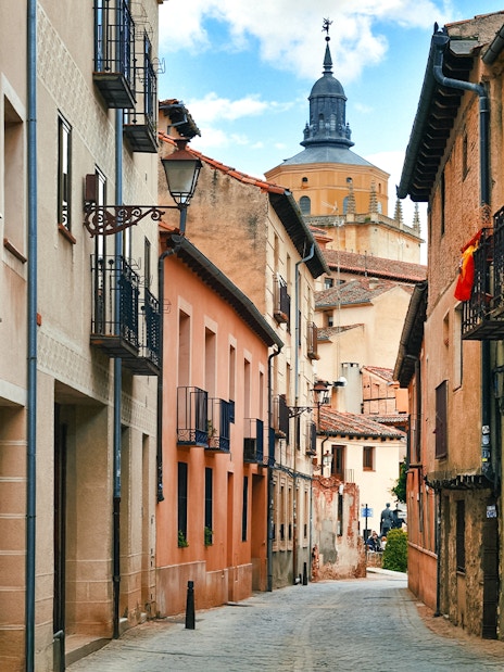 Medieval street in Segovia with historic buildings and a distant church tower.