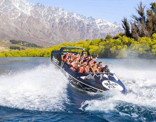 Jet boat with passengers on a river in Queenstown, surrounded by mountains and trees.