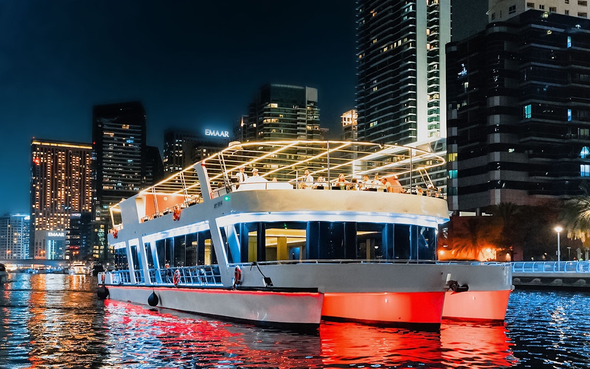 Sunset cruise boat with diners in Dubai Marina, skyscrapers illuminated in the background.