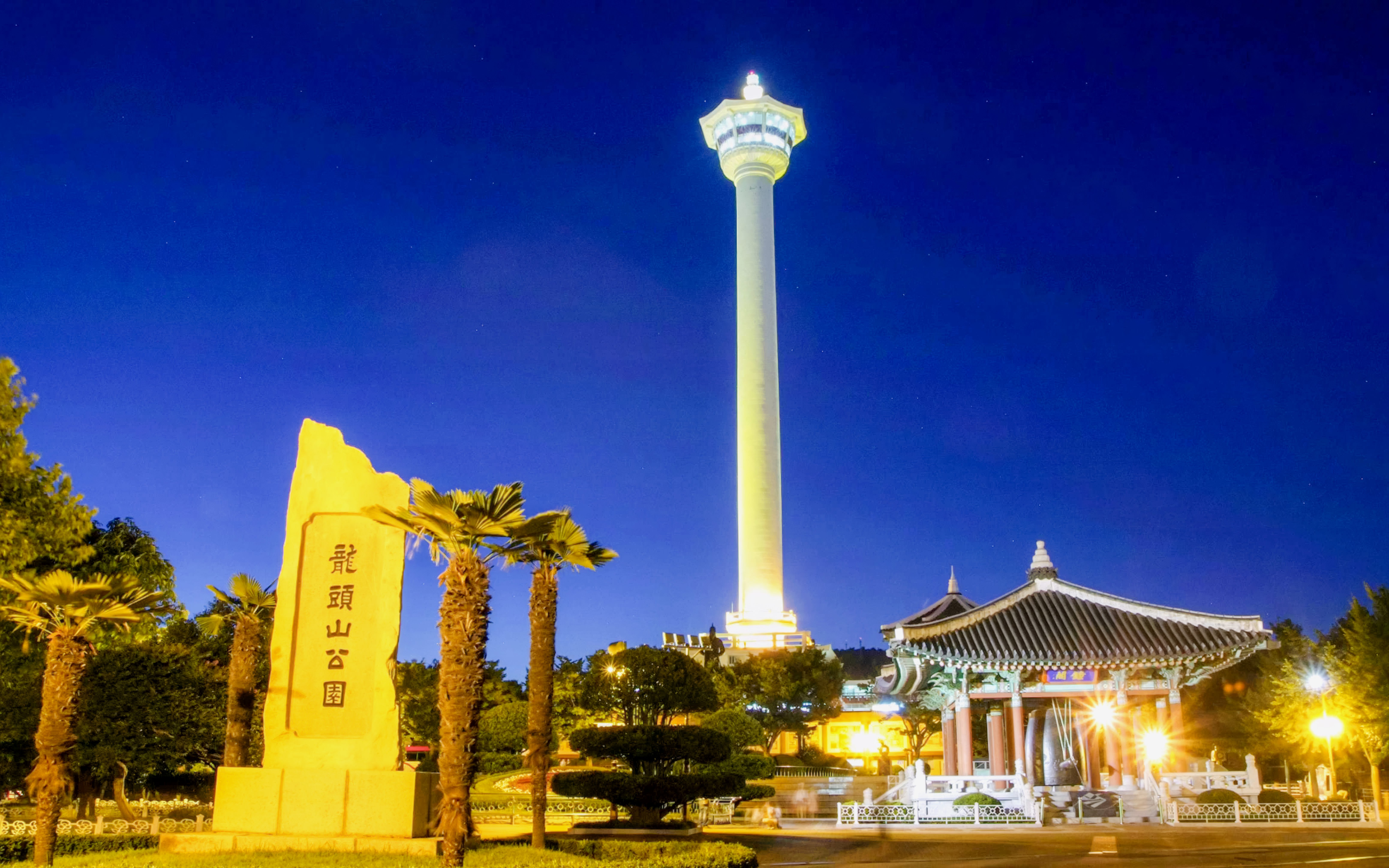 Busan Tower illuminated at night with traditional pavilion and stone monument in foreground.