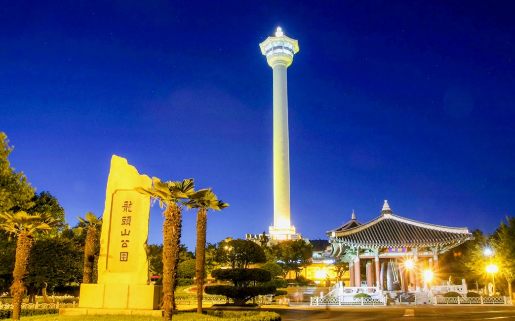 Busan Tower illuminated at night with traditional pavilion and stone monument in foreground.