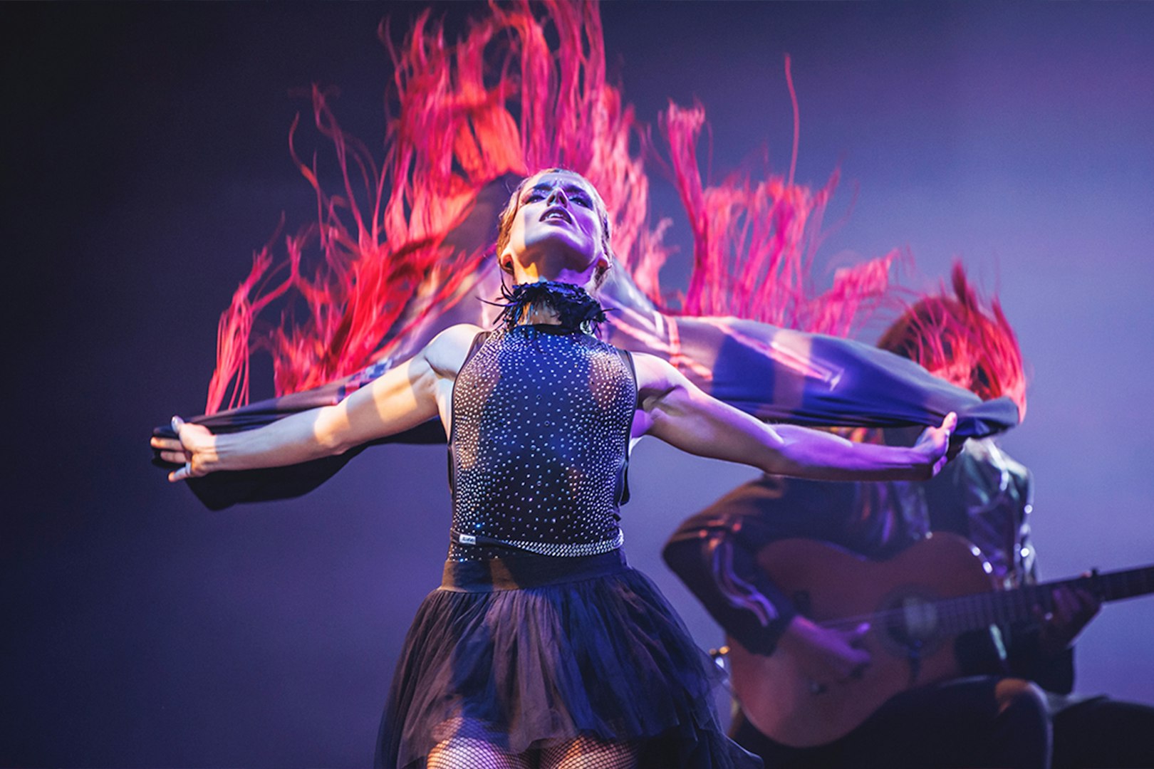Performer in dynamic dance with flowing red fabric, guitarist in background, WAH show.