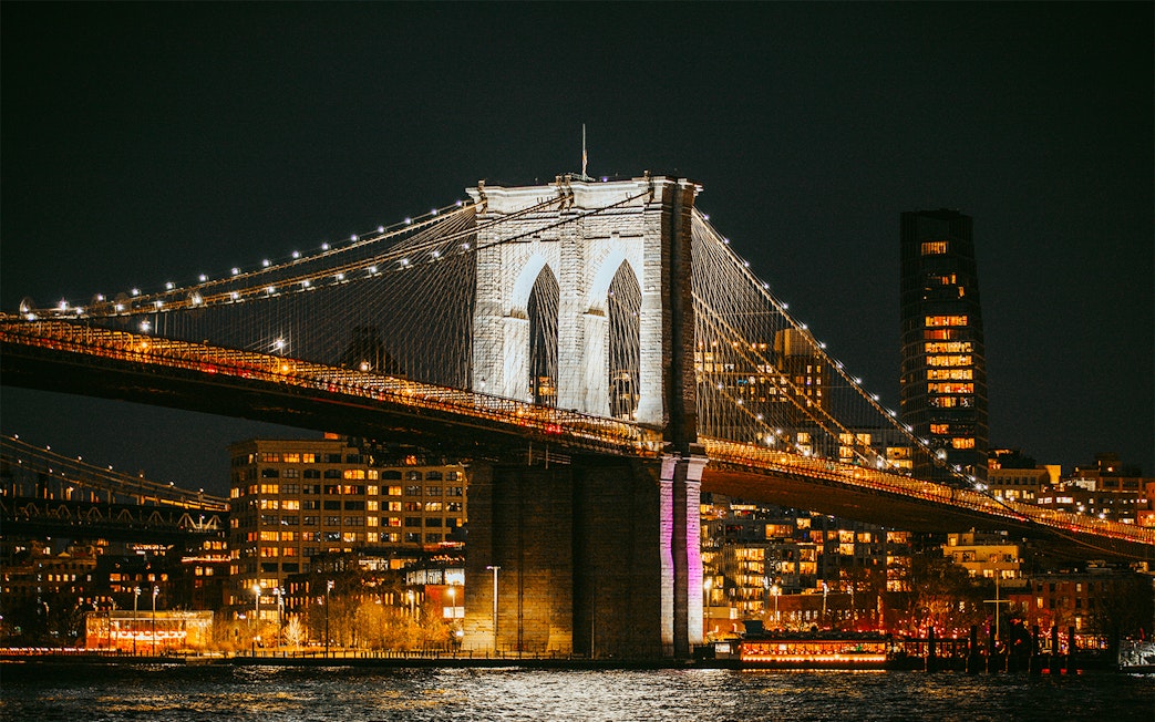 Brooklyn Bridge lit up at night over East River, New York City skyline in background.