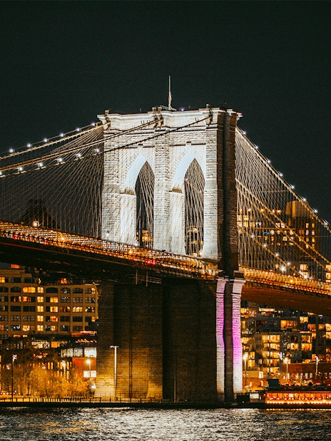 Brooklyn Bridge lit up at night over East River, New York City skyline in background.
