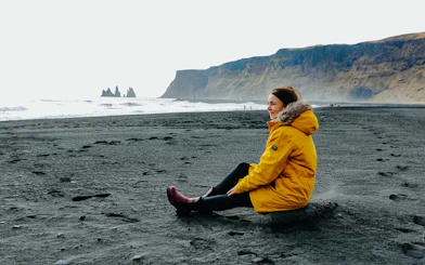 Guest sitting on Reynisfjara Black Sand Beach with sea stacks in the background, Iceland.
