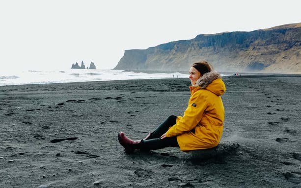 Guest sitting on Reynisfjara Black Sand Beach with sea stacks in the background, Iceland.