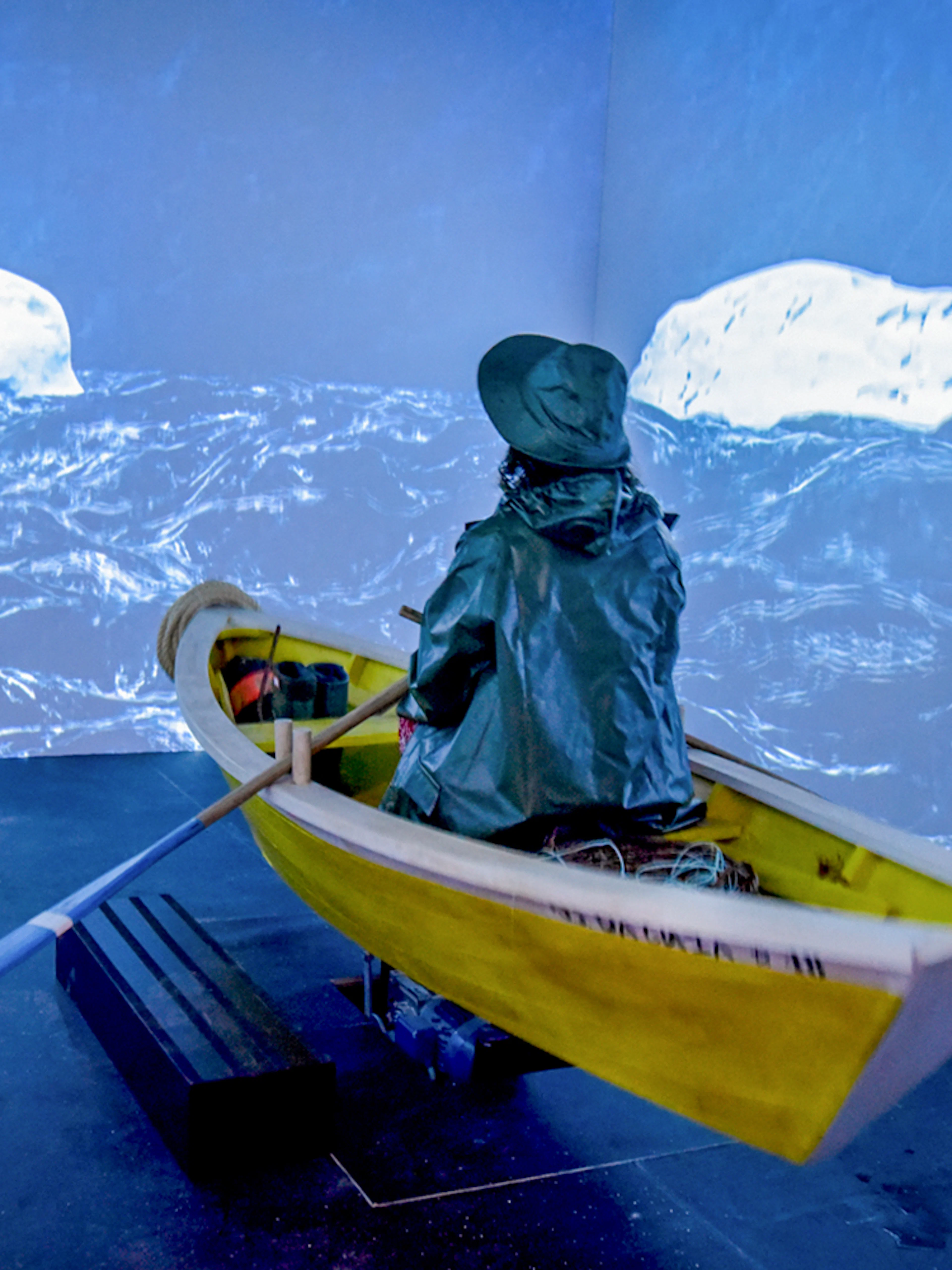 Person in raincoat rowing a yellow boat in a simulated icy sea at Codfish History Interpretation Centre.