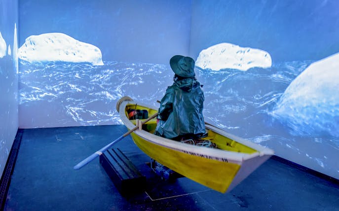 Person in raincoat rowing a yellow boat in a simulated icy sea at Codfish History Interpretation Centre.