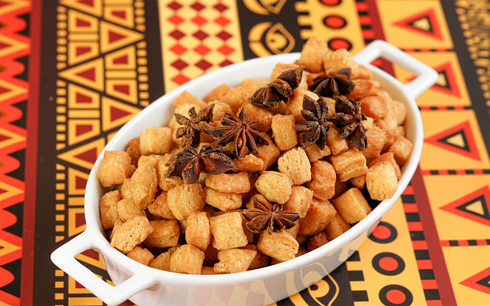 Chin Chin in a white bowl with star anise on a colorful African-patterned tablecloth.