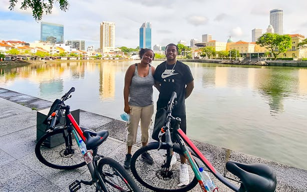 Two people with bicycles by the Singapore River on a guided historical bike tour.