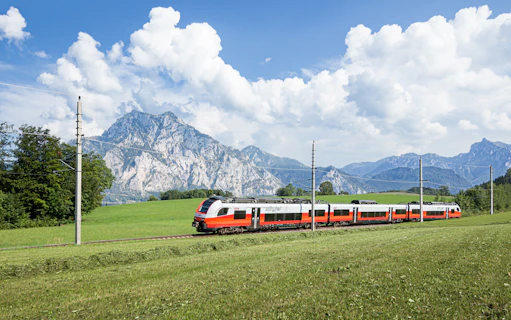 Train traveling through scenic European countryside with mountains in the background.