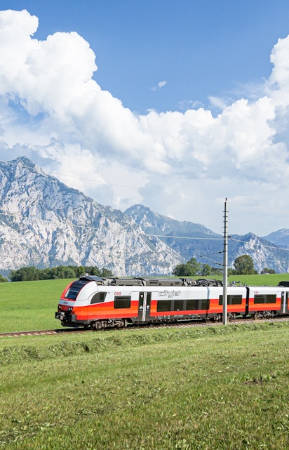 Train traveling through scenic European countryside with mountains in the background.