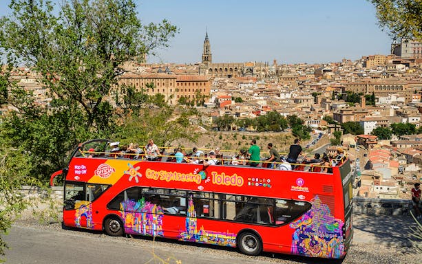 Toledo cityscape with tourists on a red hop-on-hop-off bus, featuring the Toledo Cathedral in the background.