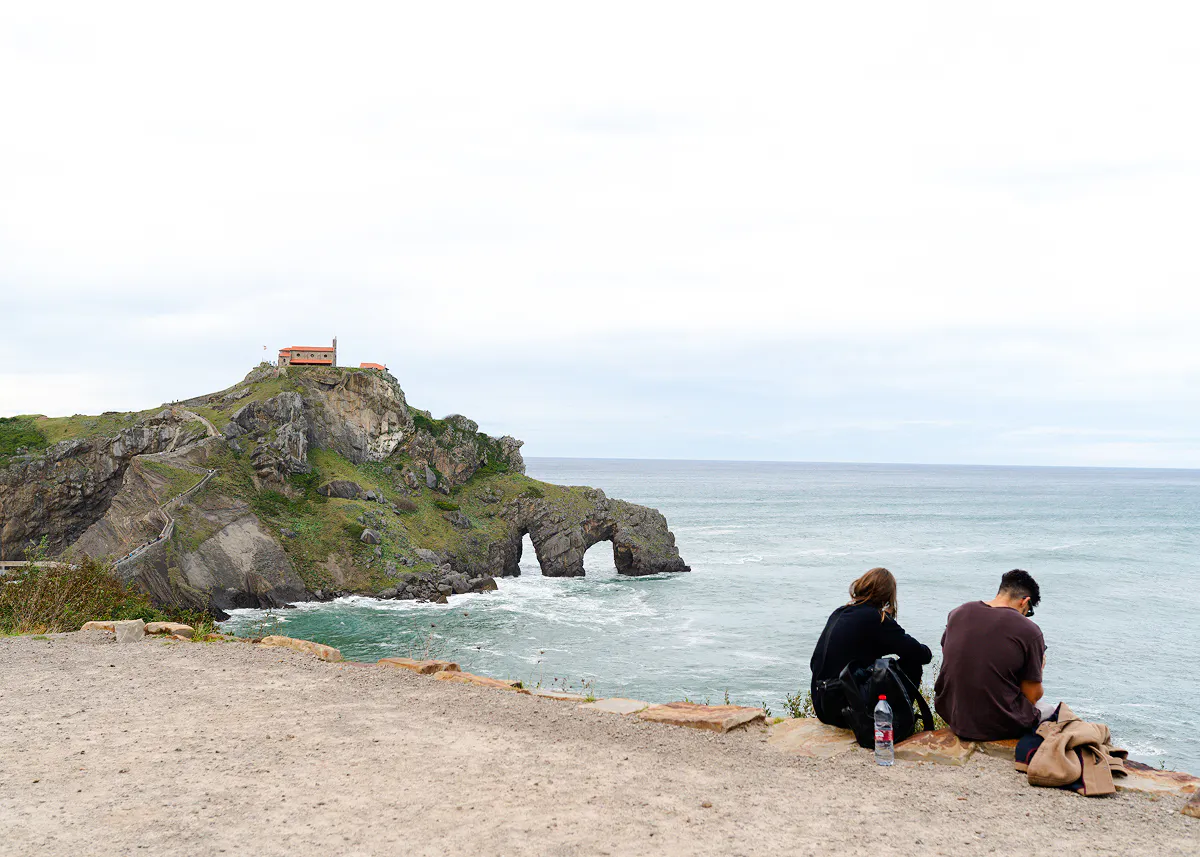 Rocky coastline with a chapel on a hill at Dragonstone, San Sebastian, with two people sitting by the sea.