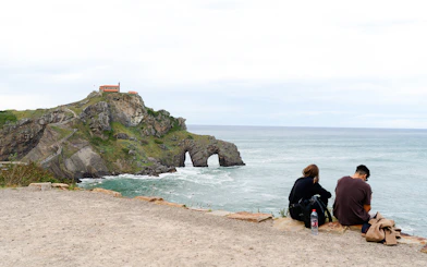 Rocky coastline with a chapel on a hill at Dragonstone, San Sebastian, with two people sitting by the sea.