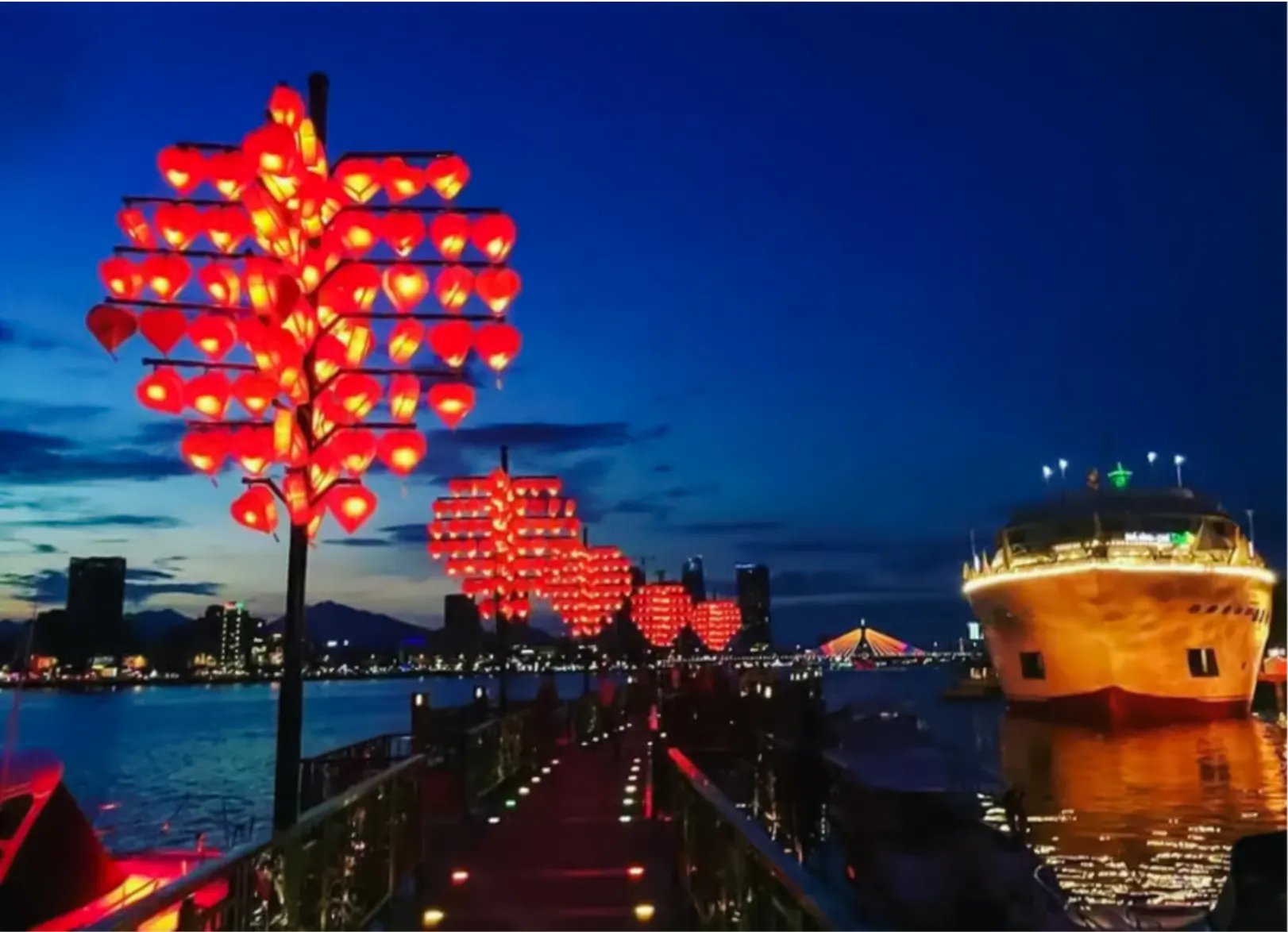 Lantern-lit walkway along Da Nang river with illuminated bridge and boat at night.