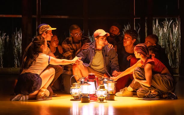 Group gathered around lanterns in a dimly lit setting, Illinoise tour experience.