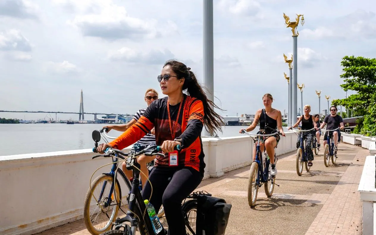 Cyclists riding along a riverside path in Bangkok with a bridge in the background.