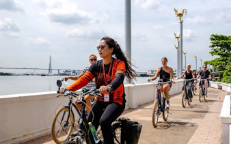 Cyclists riding along a riverside path in Bangkok with a bridge in the background.