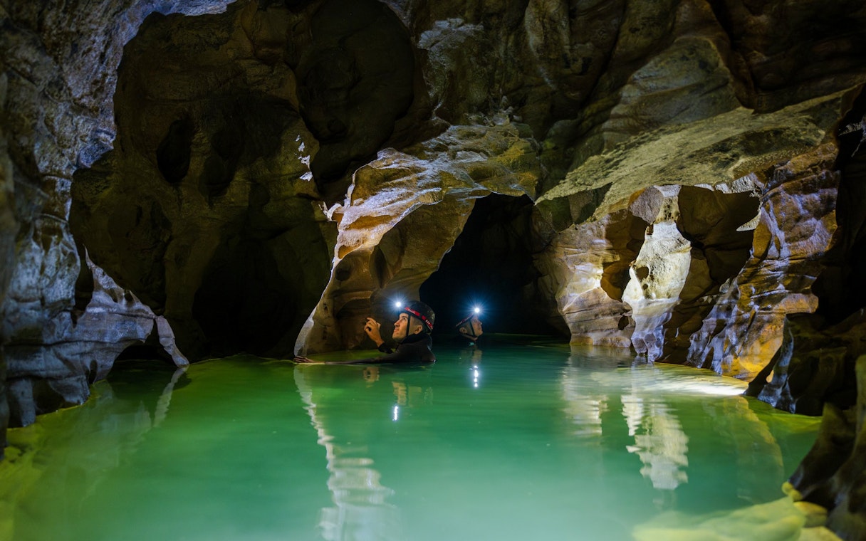 Explorers with headlamps in Okohua Glowworm Cave, Waitomo, navigating illuminated water passage.