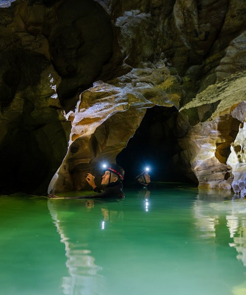 Explorers with headlamps in Okohua Glowworm Cave, Waitomo, navigating illuminated water passage.