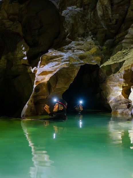 Explorers with headlamps in Okohua Glowworm Cave, Waitomo, navigating illuminated water passage.