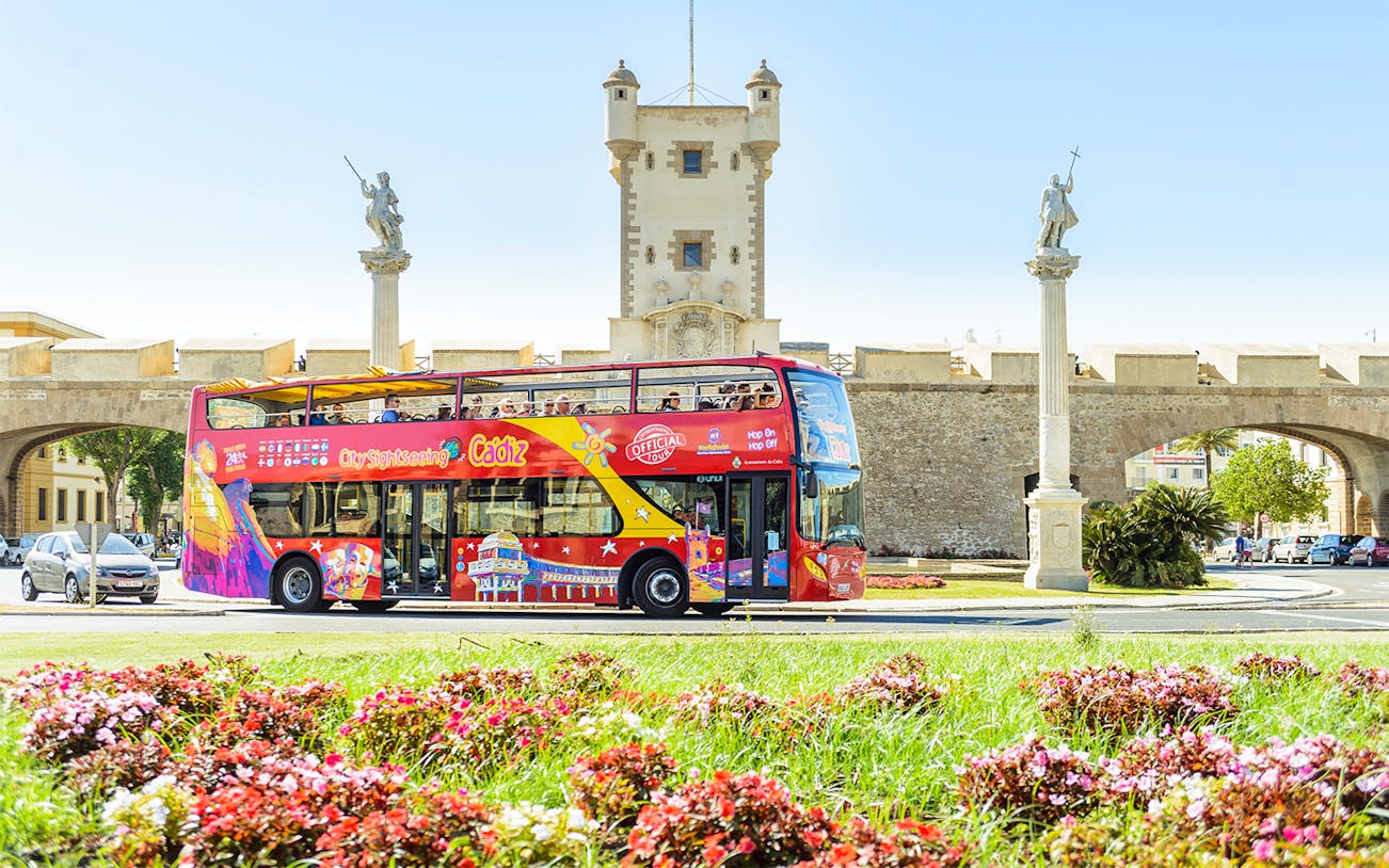 Hop-On Hop-Off Cadiz bus passing by historic city gate with statues.