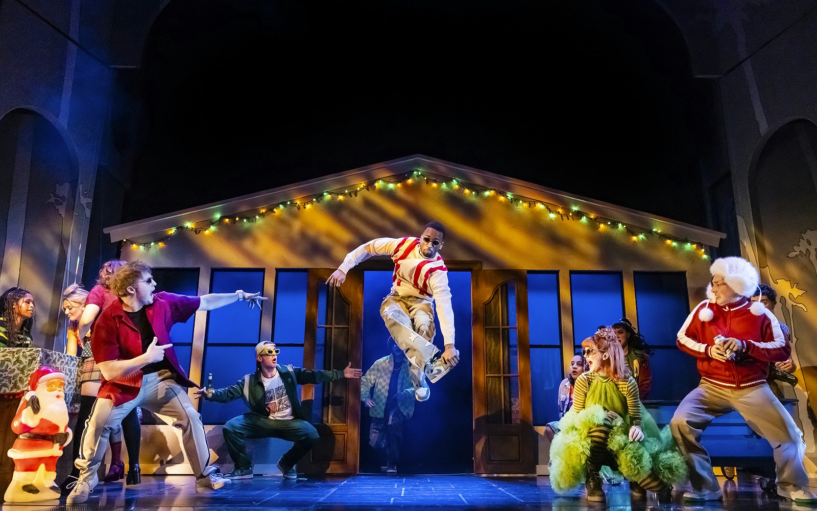 Cast performing a lively dance scene in Clueless The Musical with festive lights in the background.