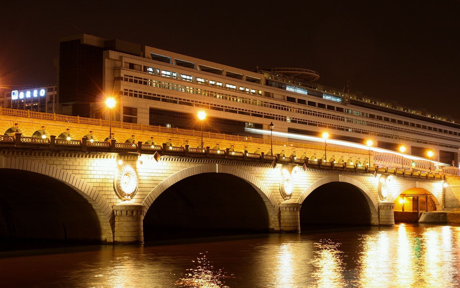 Night view of the Bibliothèque Nationale de France with illuminated bridge over the Seine River.