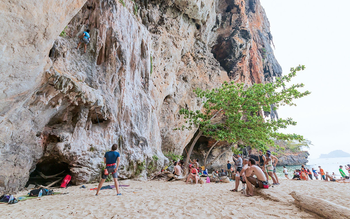 Rock climbers at Railay Beach, Krabi, scaling limestone cliffs with onlookers nearby.