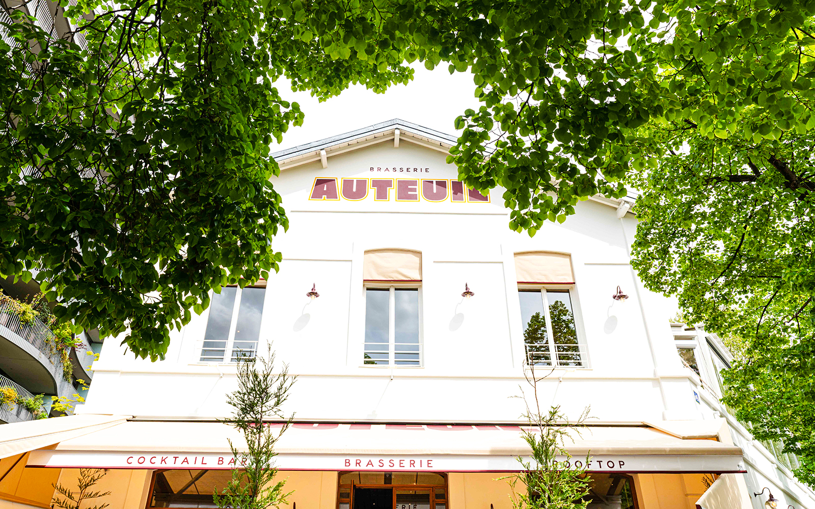 Brasserie Auteuil exterior with rooftop and cocktail bar signage, surrounded by green trees.