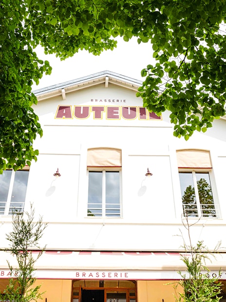 Brasserie Auteuil exterior with rooftop and cocktail bar signage, surrounded by green trees.