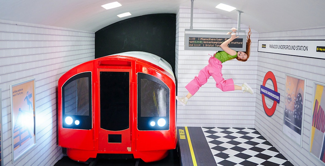 Guest hanging from a sign at Paradox Museum London with a red train in the background.