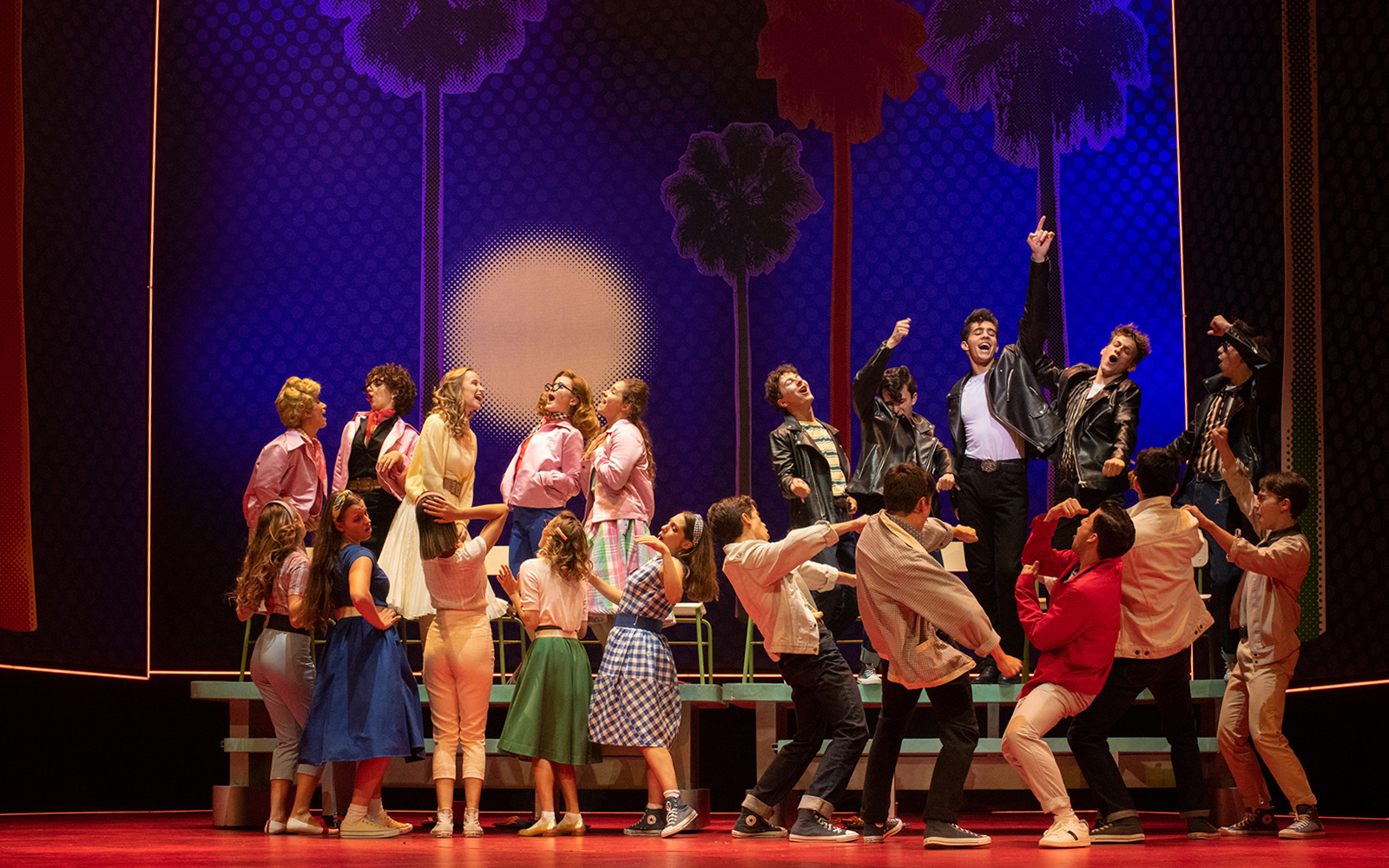 Cast performing a lively scene from the musical "Grease" with colorful costumes and dynamic choreography.