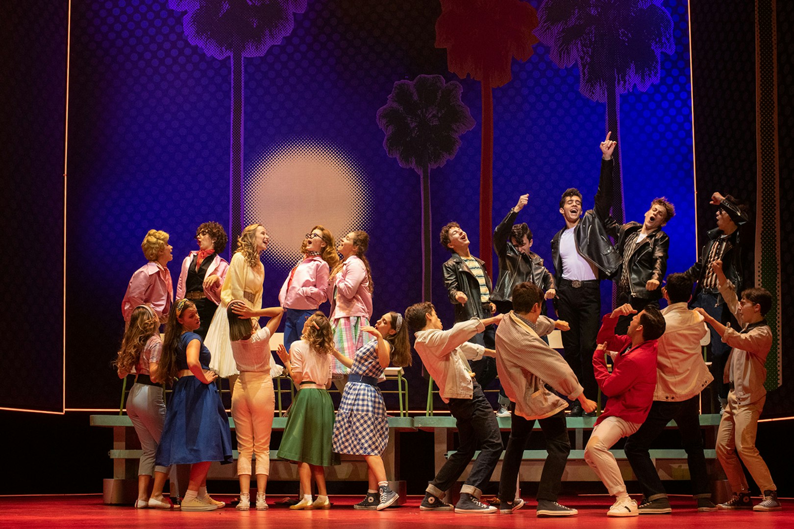 Cast performing a lively scene from the musical "Grease" with colorful costumes and dynamic choreography.
