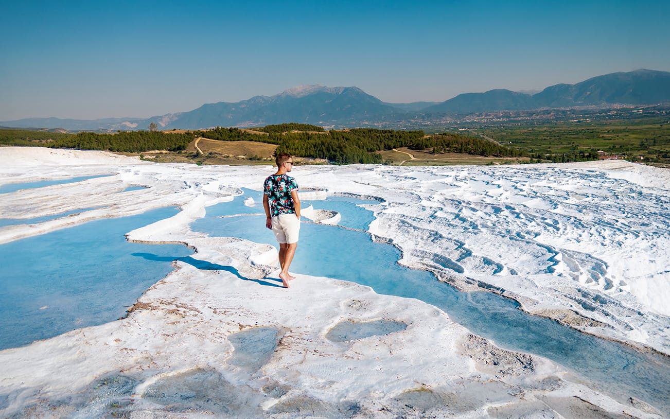 Person walking on travertine terraces at Pamukkale, Turkey, with mountains in the background.