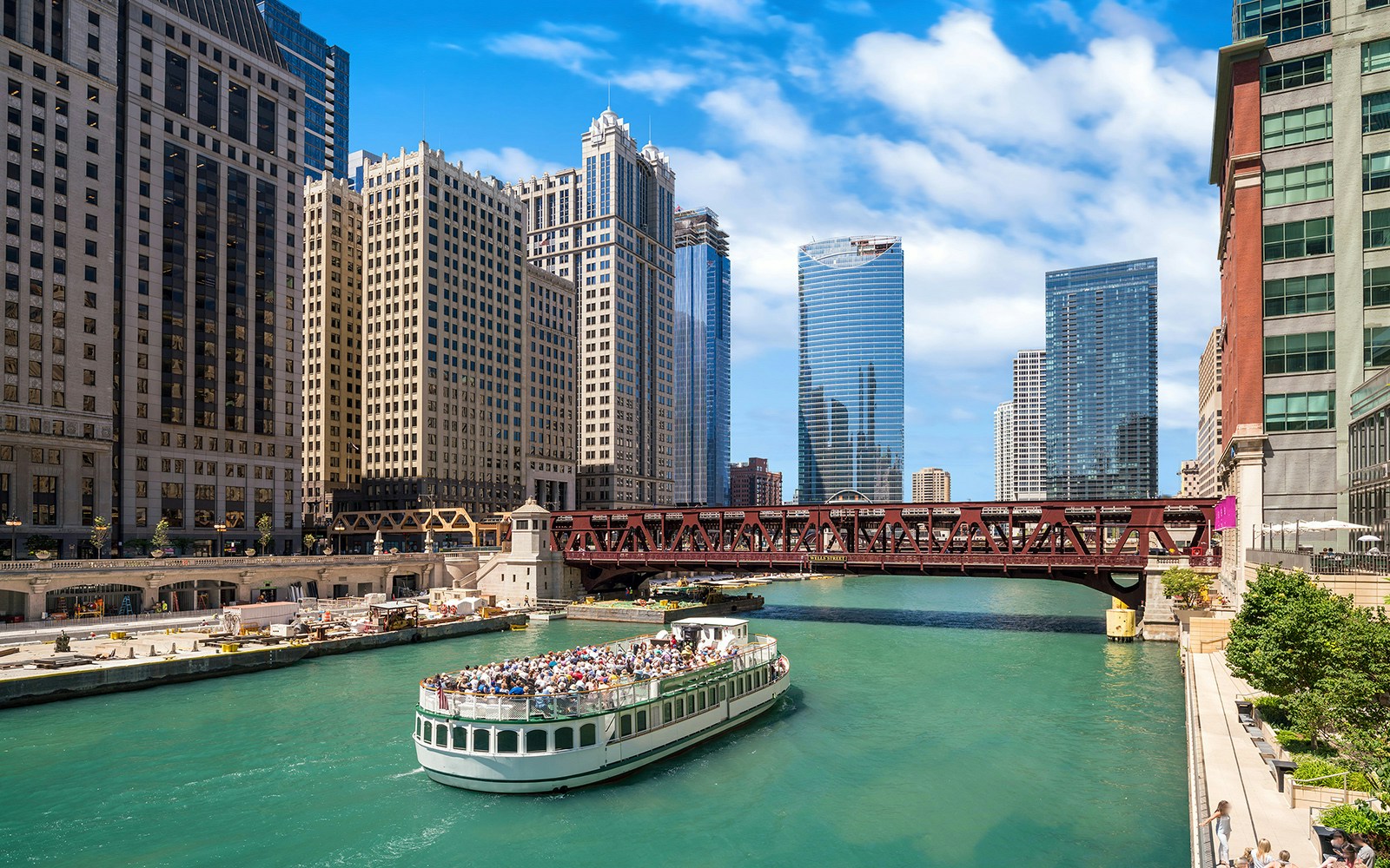 Chicago River cruise boat passing under a bridge with city skyscrapers in the background.