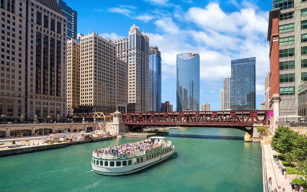 Chicago River cruise boat passing under a bridge with city skyscrapers in the background.